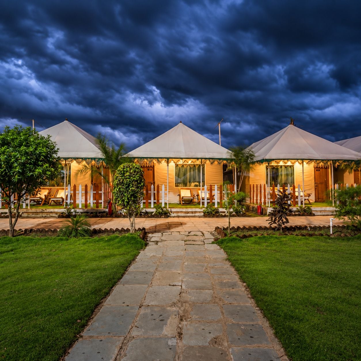 A row of three tents with a stone pathway leading to them, surrounded by grass and trees.