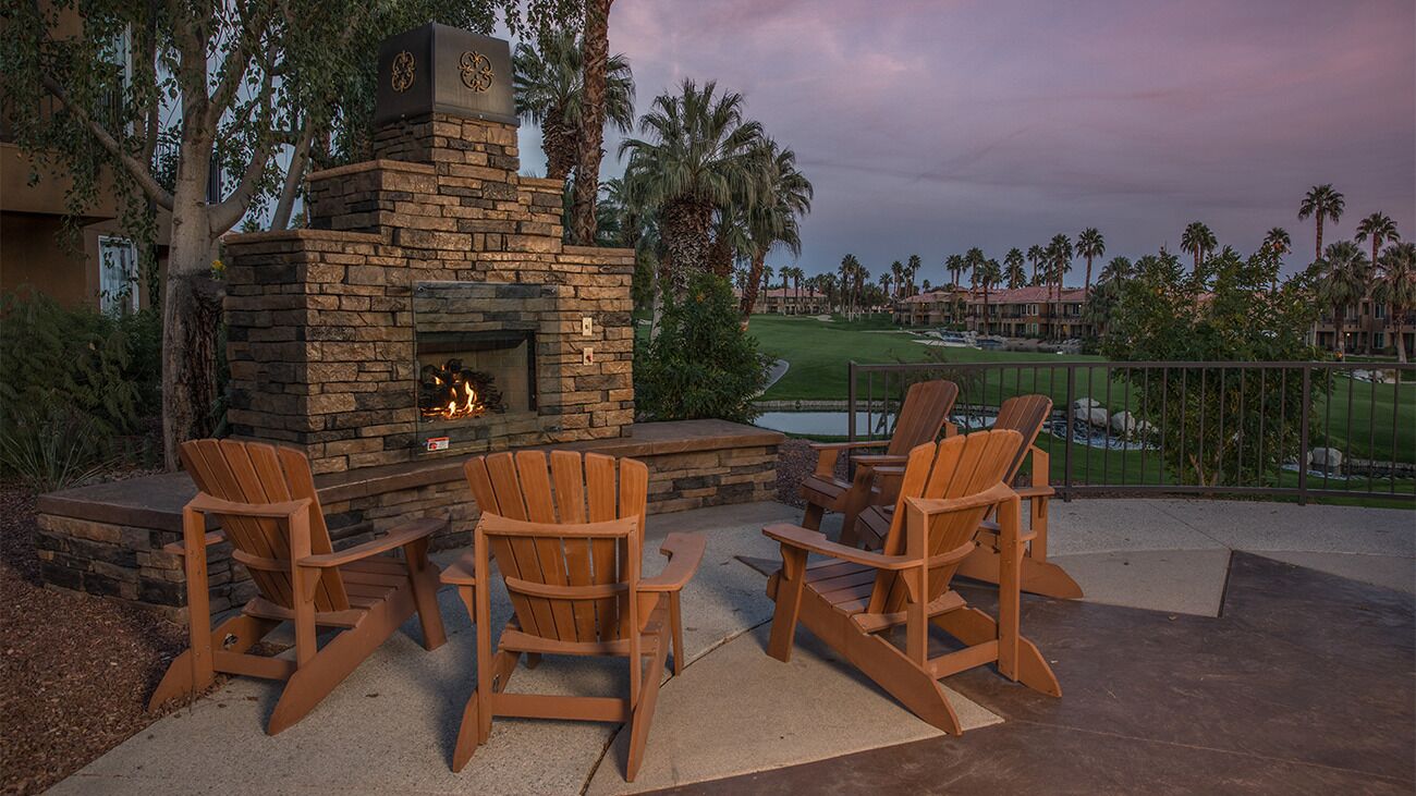 Une cheminée en pierre avec un feu de bois, entourée de fauteuils Adirondack en bois, installée sur une terrasse offrant une vue sur un terrain de golf et une piscine.