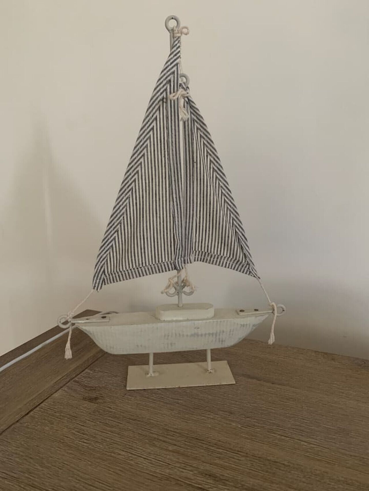 Een modelzeilboot met een gestreepte zeil en een klein pronkstuk, geplaatst op een houten tafel voor een effen muur.
