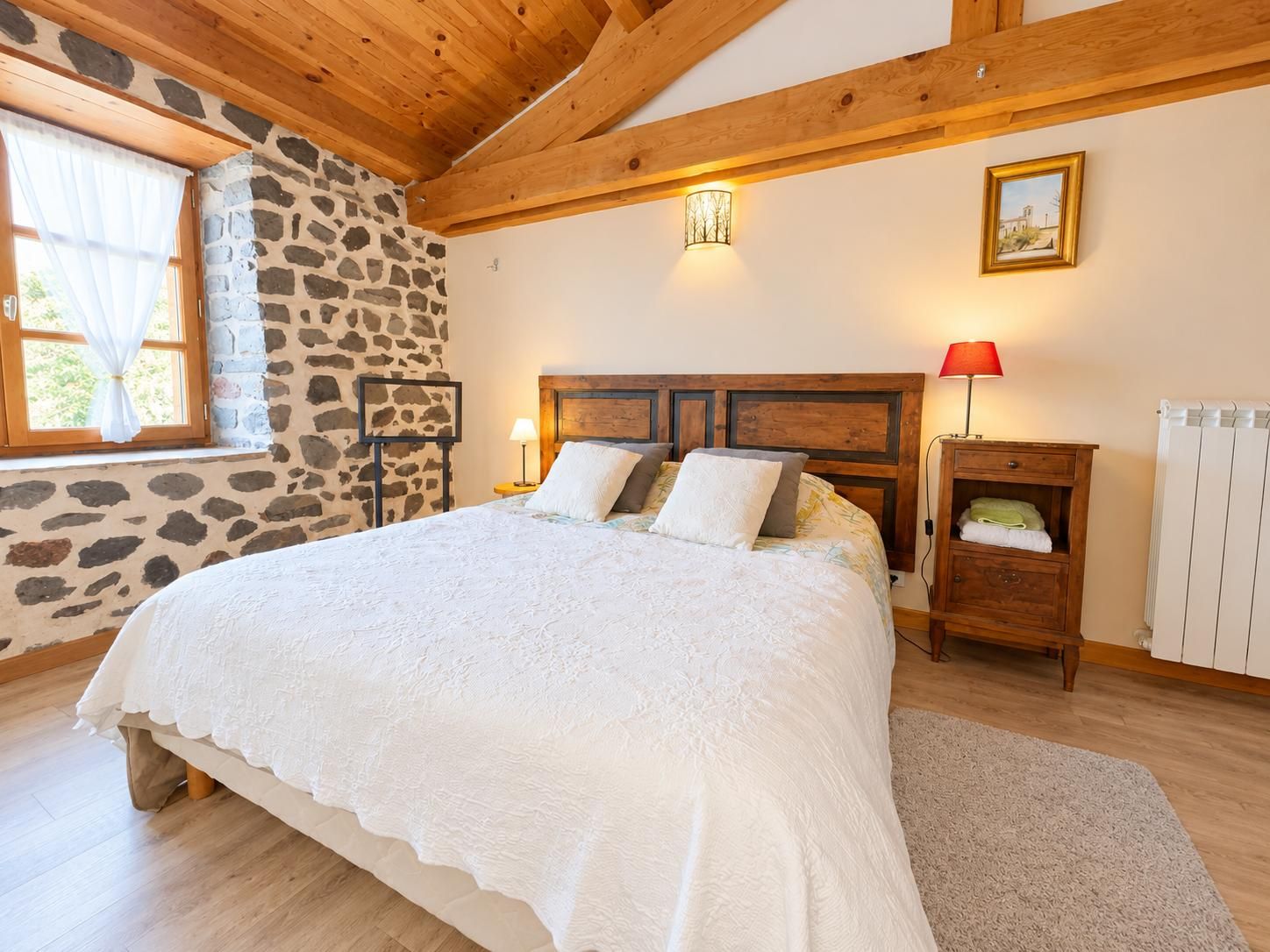 Une chambre avec un mur en pierre, des poutres apparentes, un lit recouvert d’une courtepointe blanche, une table de chevet avec une lampe et un radiateur.