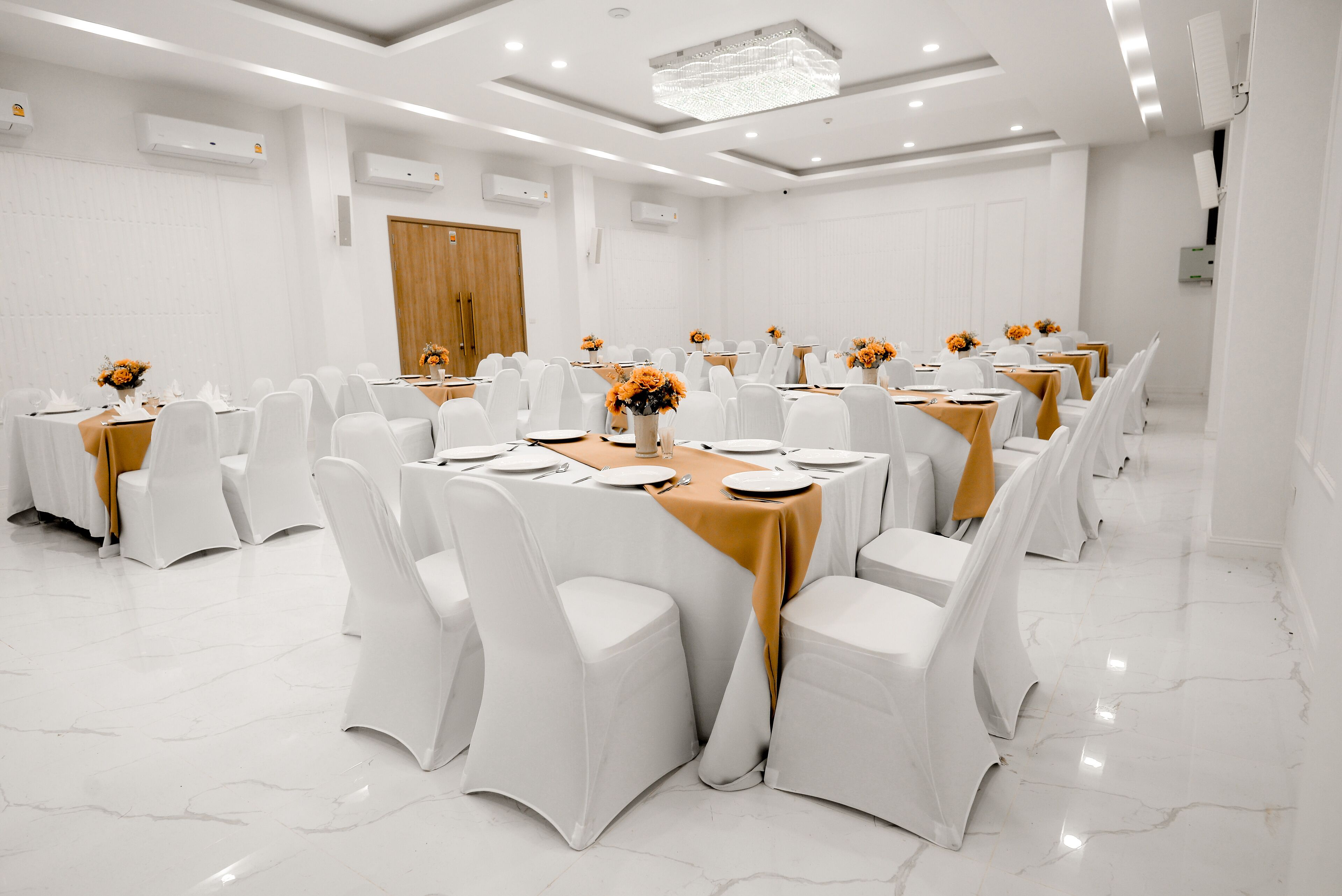 A conference room with white chairs, tables with orange tablecloths, and flower centerpieces.