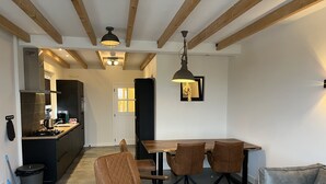 A modern kitchen with dark cabinetry, a gas stove, and a coffee machine. There is a dining area with a wooden table and four brown chairs. A gray sofa is in the foreground. - Christinahoeve Apartment #8 (Boskoop)