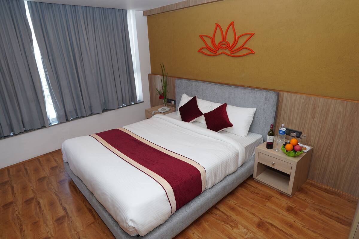 A neatly made bed with a red and white bedspread, a wooden nightstand with a bottle of wine, a glass of water, and a bowl of fruit.