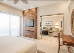 Quarto de hotel com uma cama grande, uma televisão fixada numa parede texturizada, um espaço de refeições com mesa e cadeiras, e vista para a área de estar adjacente.