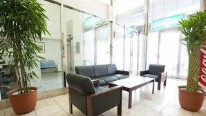 A modern waiting area with a sofa, chairs, and a coffee table. There are potted plants and a glass wall with a view of the outside. - Hotel Yassa (Mihara)