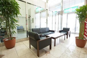 Un área de espera moderna con un sofá, sillas y una mesita de centro. Hay plantas en macetas y un muro de cristal con vistas al exterior.