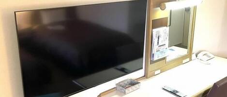 A hotel room with a flat-screen TV mounted on the wall, a vanity with a mirror and a telephone, and a desk with a chair.