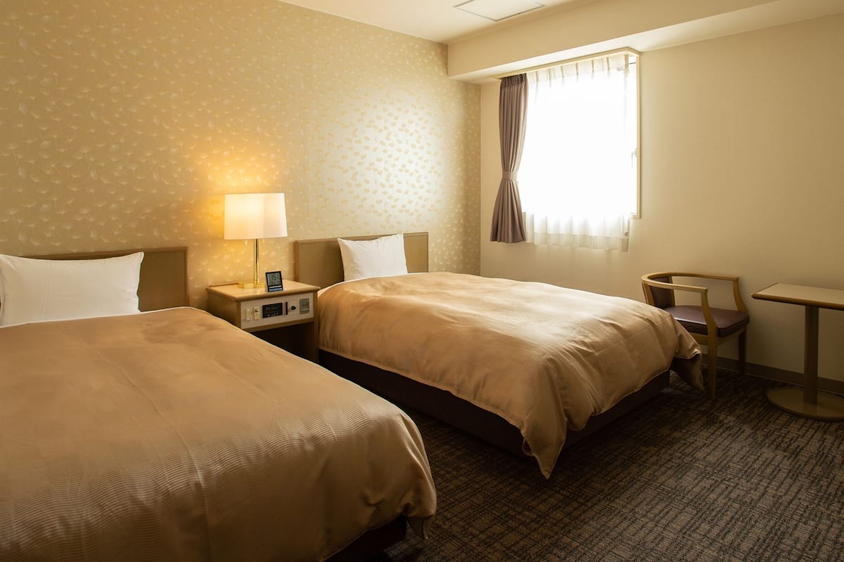 A hotel room with two beds, a nightstand with a lamp, a small table, and a window with curtains.