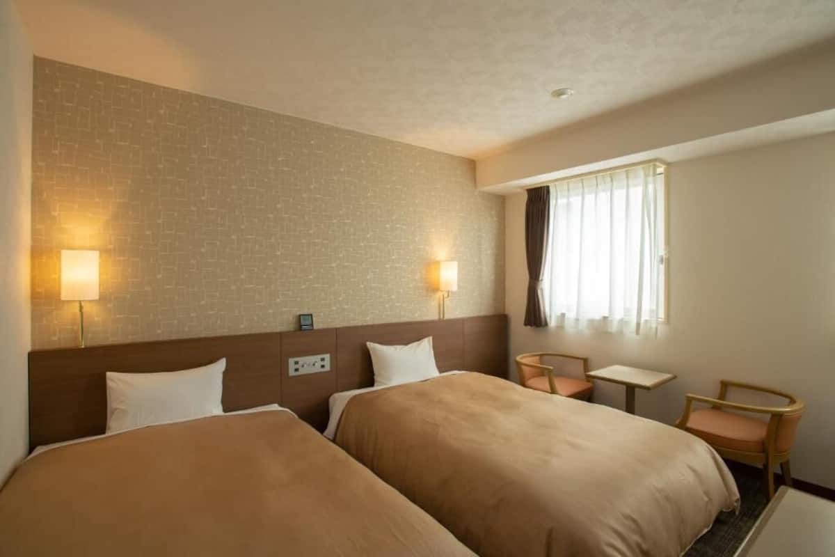 A hotel room with two beds, a small table, a chair, and a window with curtains.