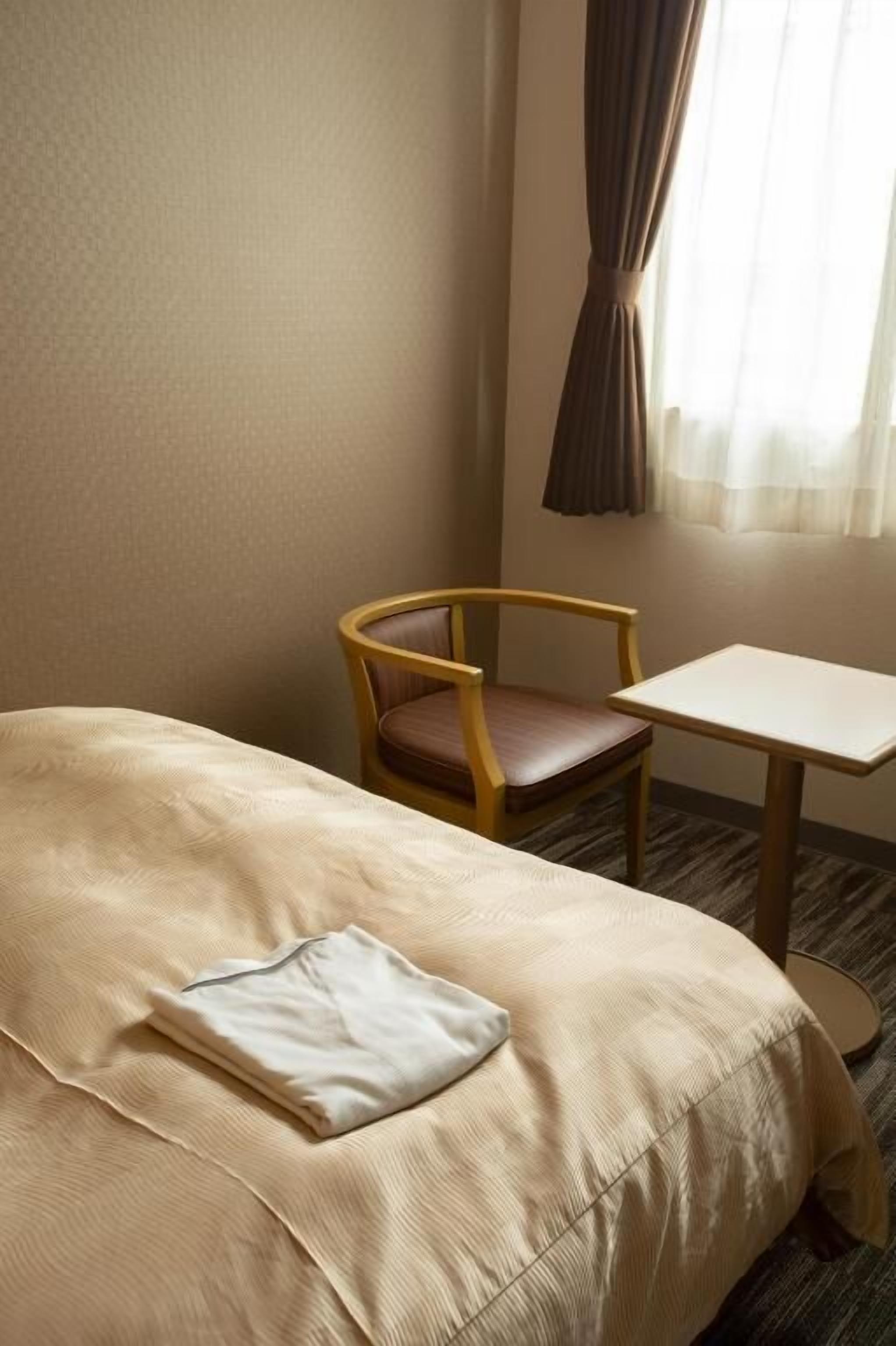 A hotel room with a bed, a wooden chair, a small table, and a window with curtains.