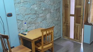 A corner room with a stone wall, a wooden door, a small table with a chair, and a bed with a wooden headboard.
