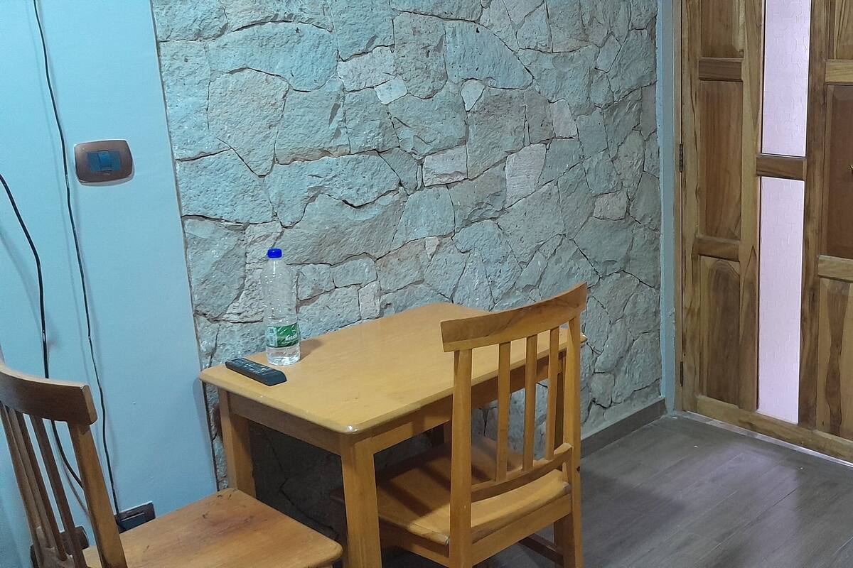 A corner room with a stone wall, a wooden door, a small table with a chair, and a bed with a wooden headboard.