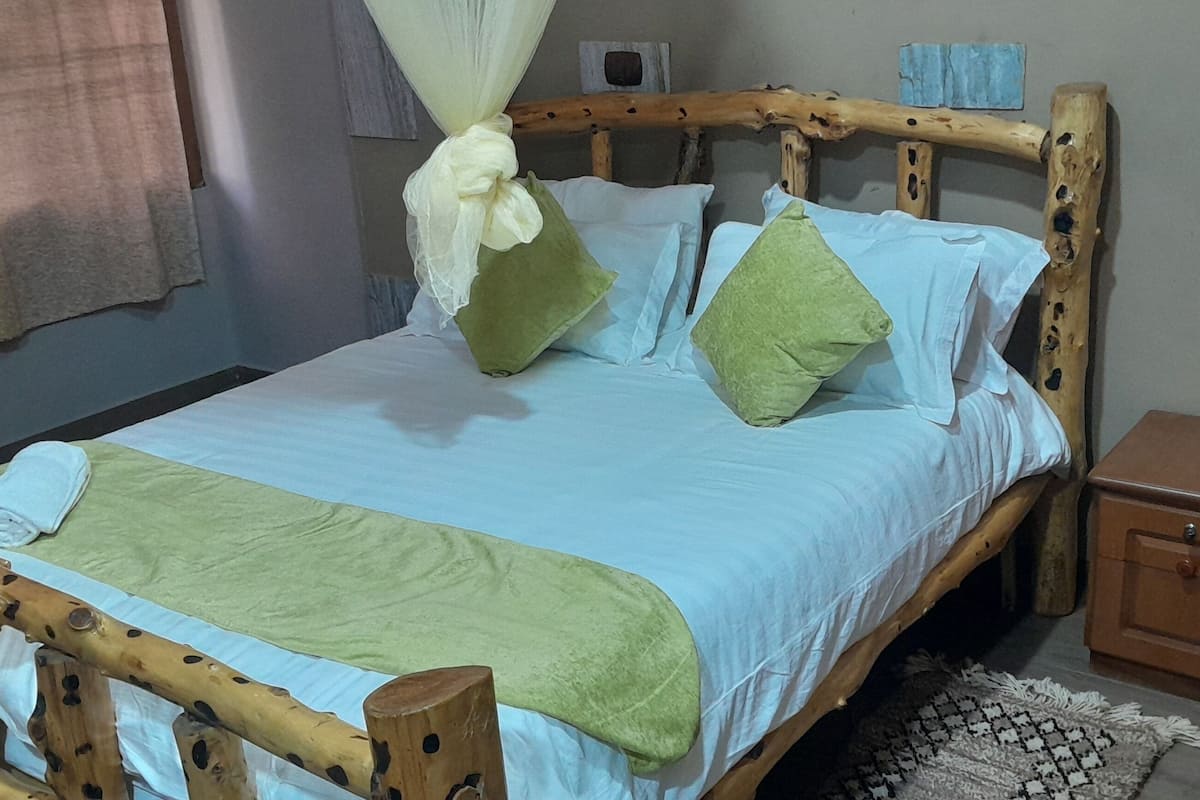 A bed with a wooden frame, a green bedspread, and pillows. There is a nightstand with a drawer and a small rug on the floor.