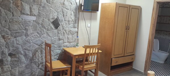 Una habitación pequeña con pared de piedra, un armario de madera, una mesa de madera con dos sillas y un inodoro con suelo estampado.