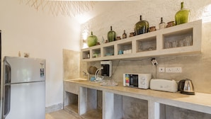 A kitchen with a built-in refrigerator, a microwave, a toaster, and a coffee maker on the counter. There are shelves above the counter holding various glassware and bottles. - Beach front villa Myconos (Anjuna)