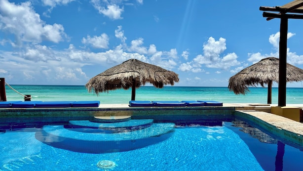 Una piscina azul cristalina con dos sombrillas de paja y sillas de descanso azules.