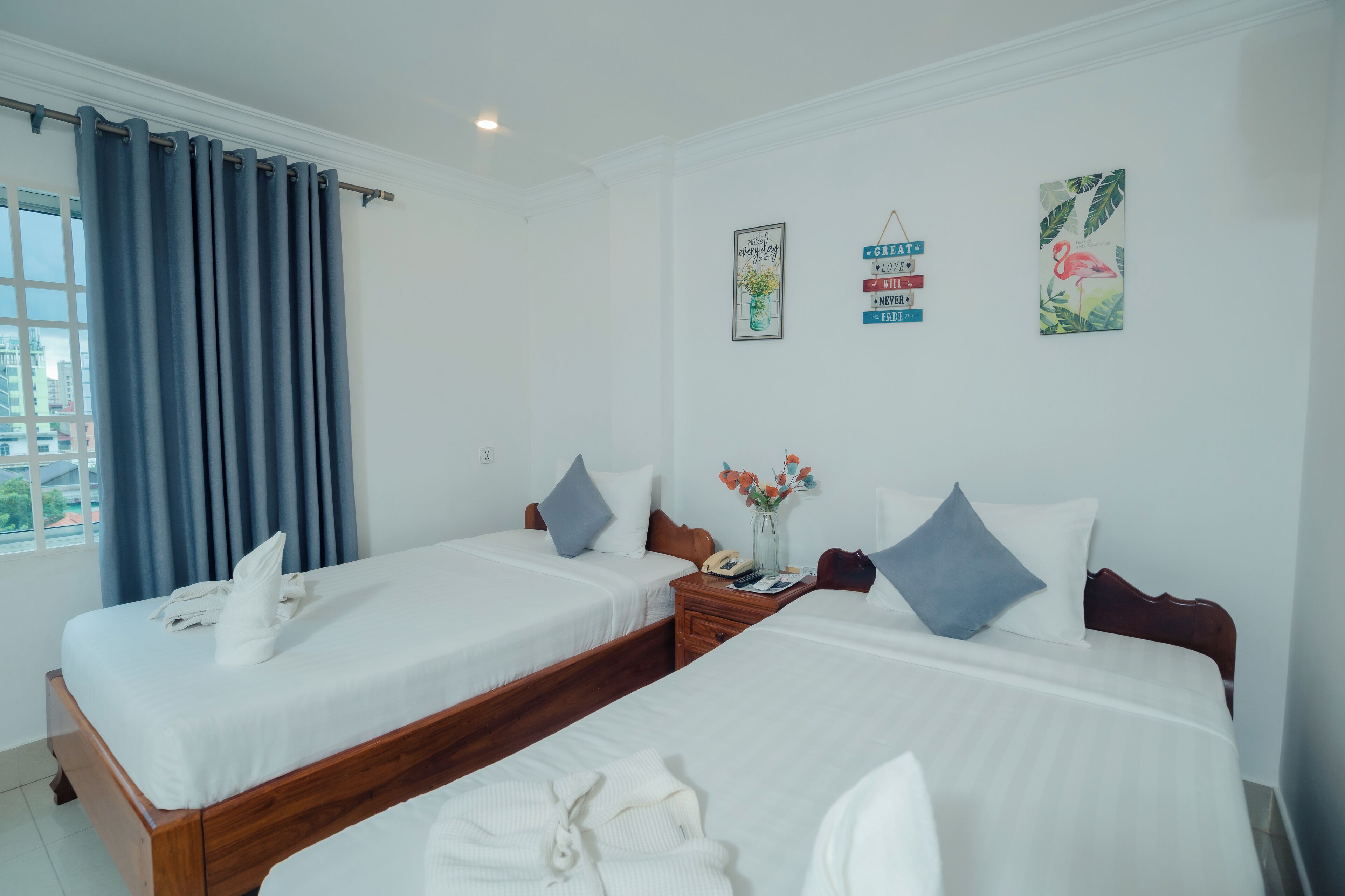A hotel room with two beds, a wooden headboard, a nightstand with a phone, a vase with flowers, and wall art.
