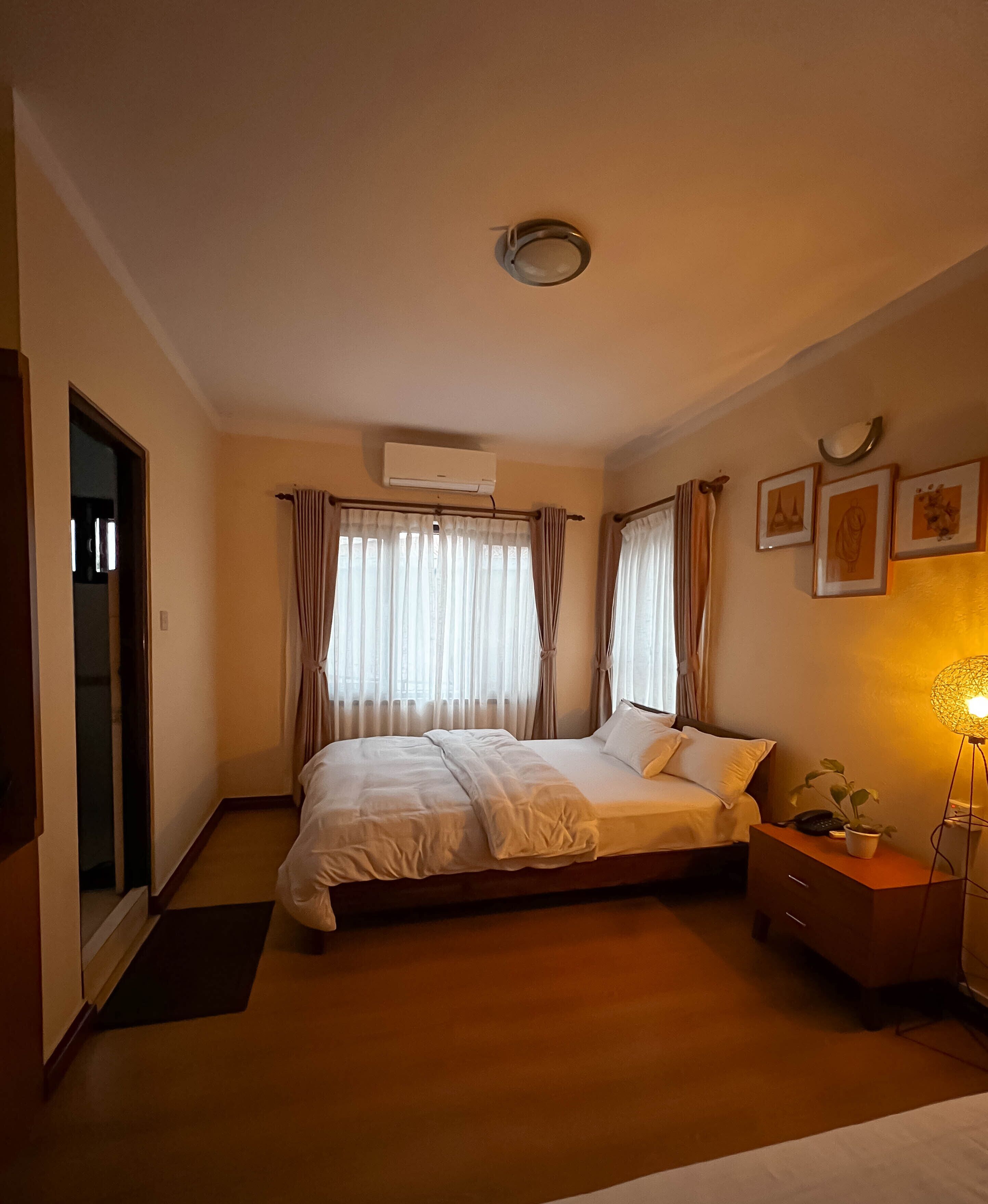 A hotel room with a bed, bedside table, lamp, air conditioning unit, curtains, and framed pictures on the wall.