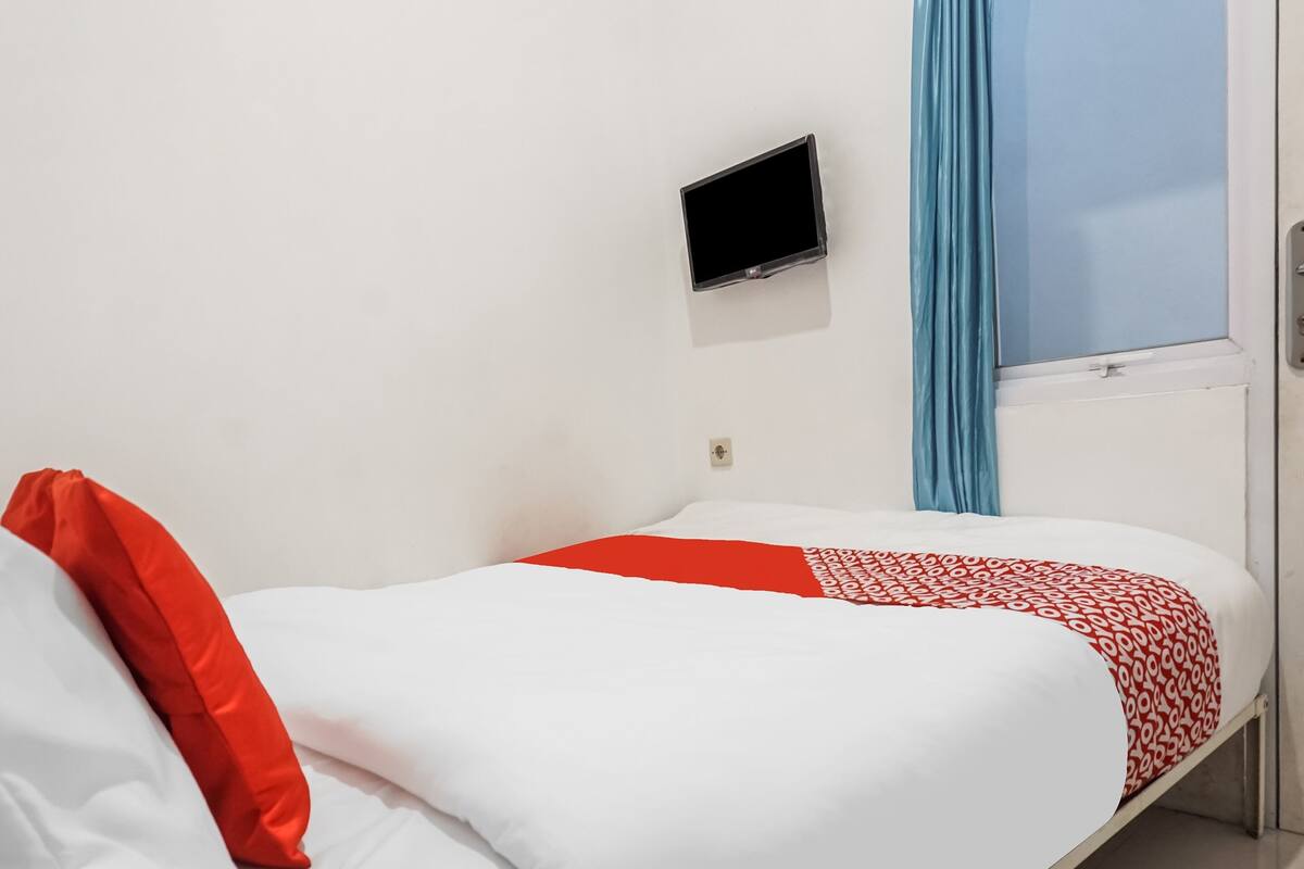 Ein Einzelbett mit weißroter Bettdecke, einem roten Kissen, einem kleinen Fernseher an der Wand und einem Fenster mit blauen Vorhängen.
