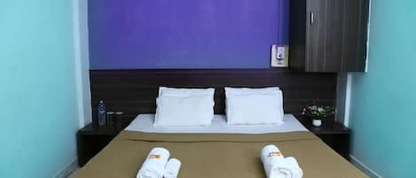 Habitación de hotel con una cama, dos almohadas, una mesita de noche con una botella y vasos, una planta pequeña en maceta y un armario.