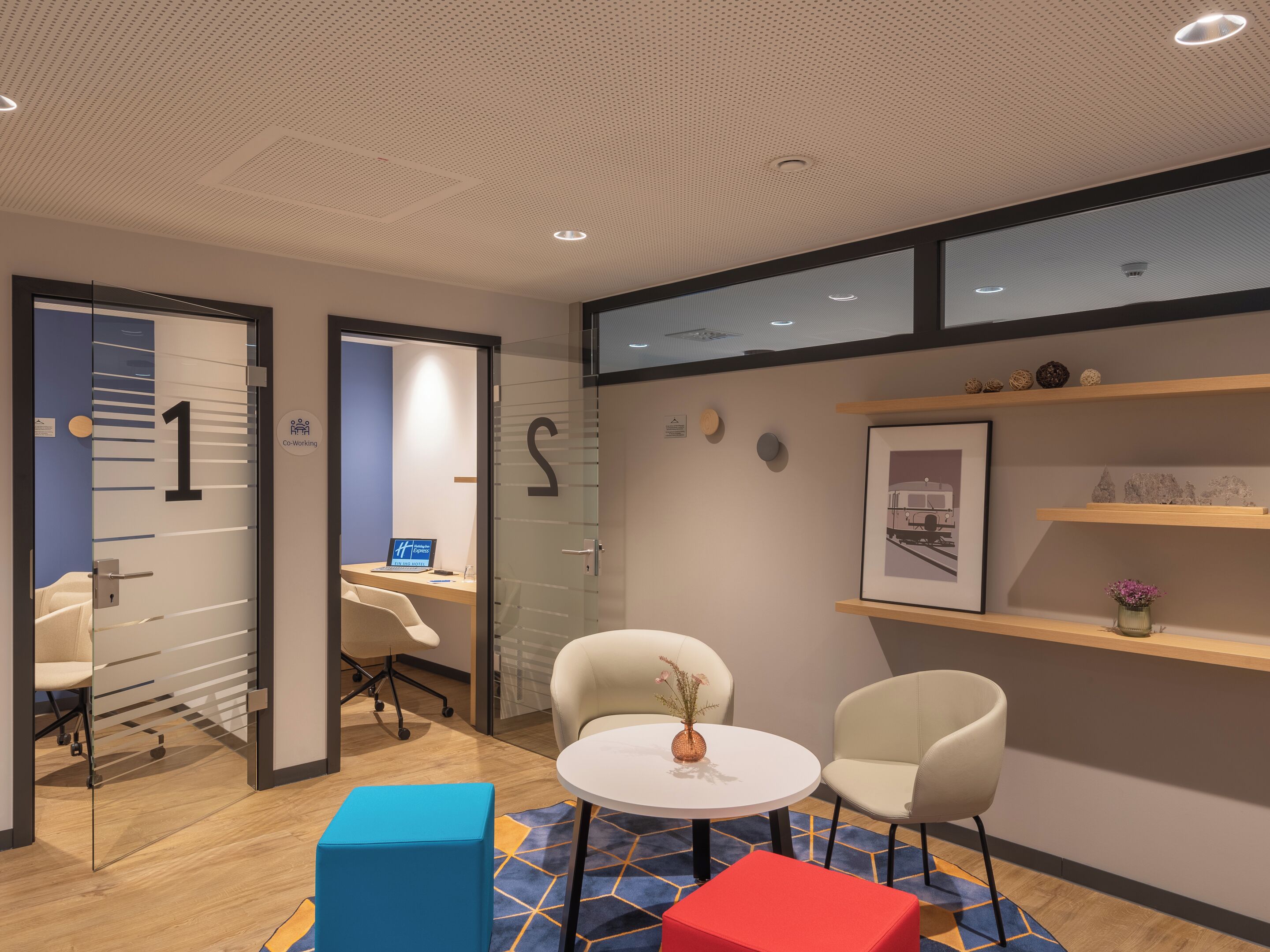 A modern office space with glass partitions, numbered doors, a round table with chairs, and a blue and red ottoman.