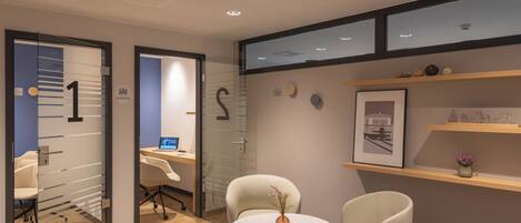 A modern office space with glass partitions, numbered doors, a round table with chairs, and a blue and red ottoman.