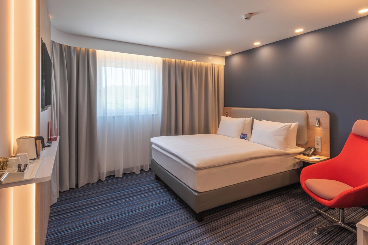 A hotel room with a bed, a red chair, a bedside table with a lamp, and a window with curtains.