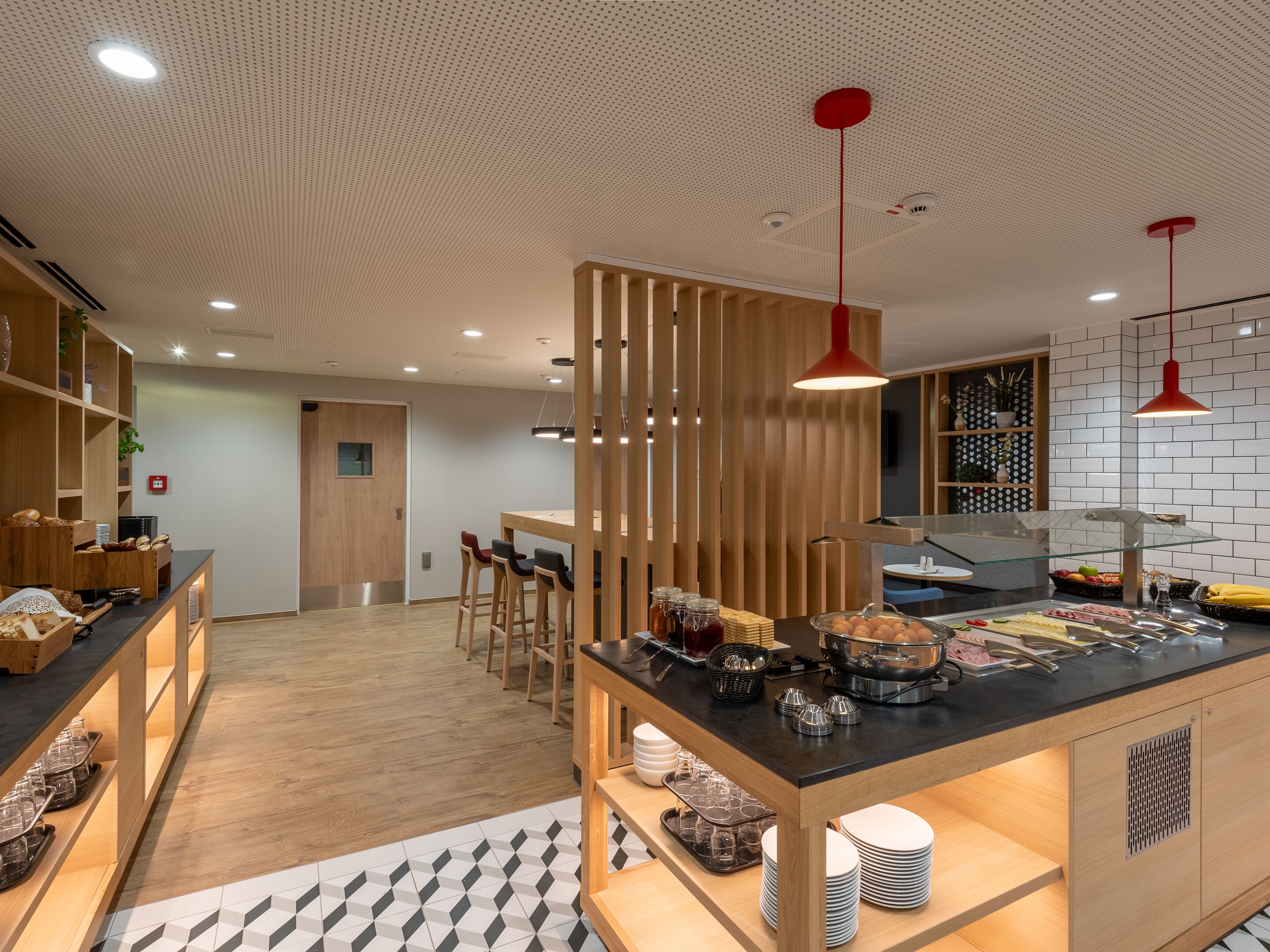 Um café moderno com móveis de madeira, balcão preto com produtos alimentícios e luminárias pendentes vermelhas.