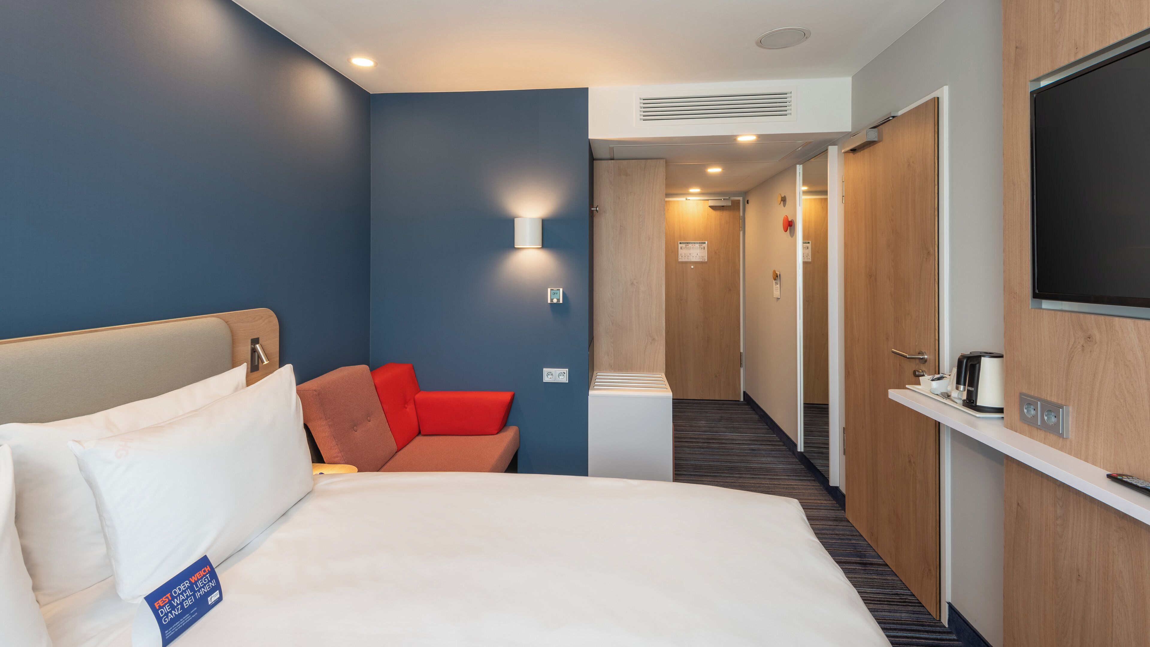Ein Hotelzimmer mit einem Bett, einer Sitzecke mit rotem Kissen, einem an der Wand montierten Fernseher und einer Wandverkleidung aus Holz.