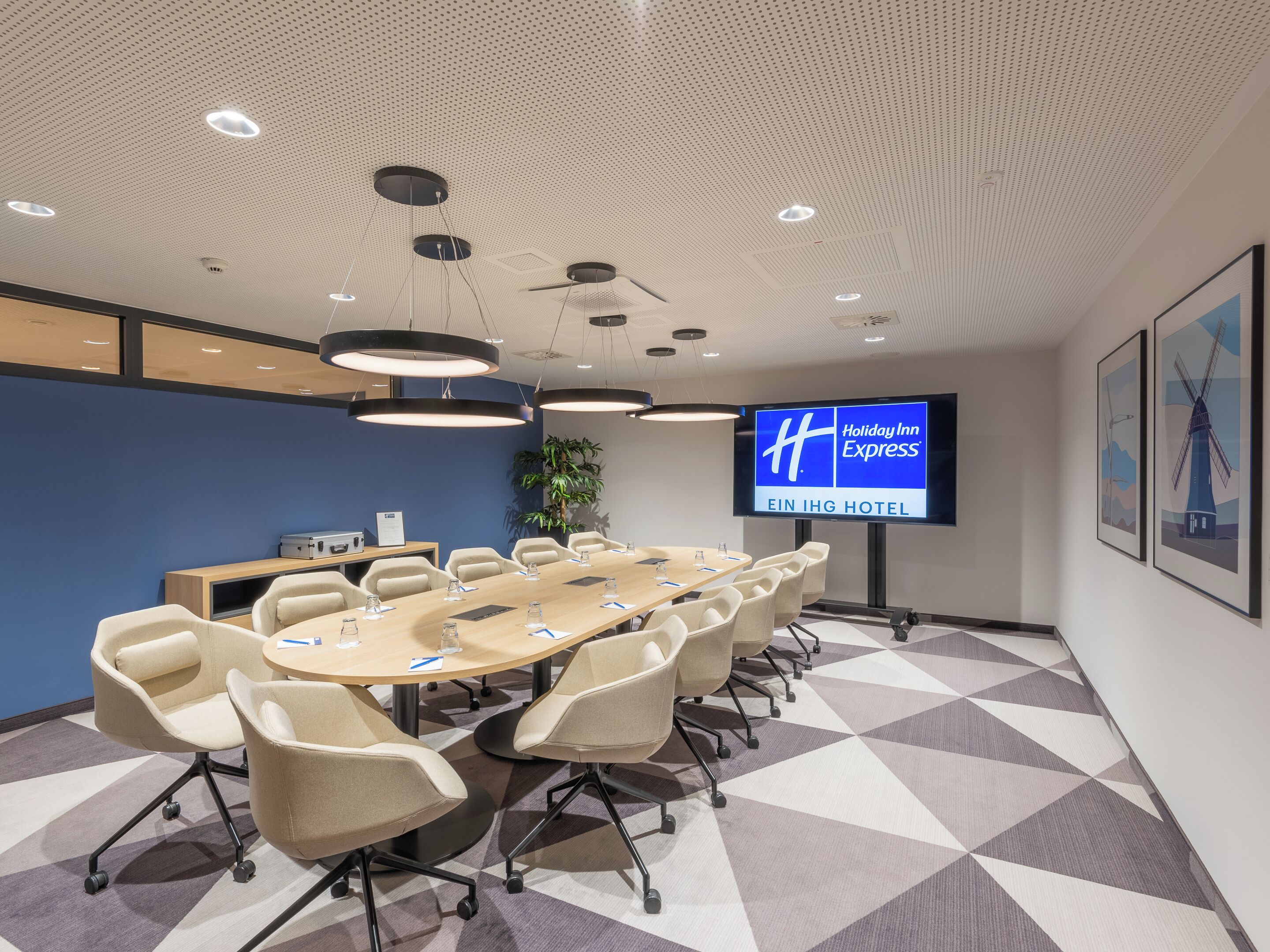 Un salón de conferencias con una mesa larga, varias sillas, una pantalla grande que muestra el logotipo de Holiday Inn Express y una alfombra con estampado geométrico.