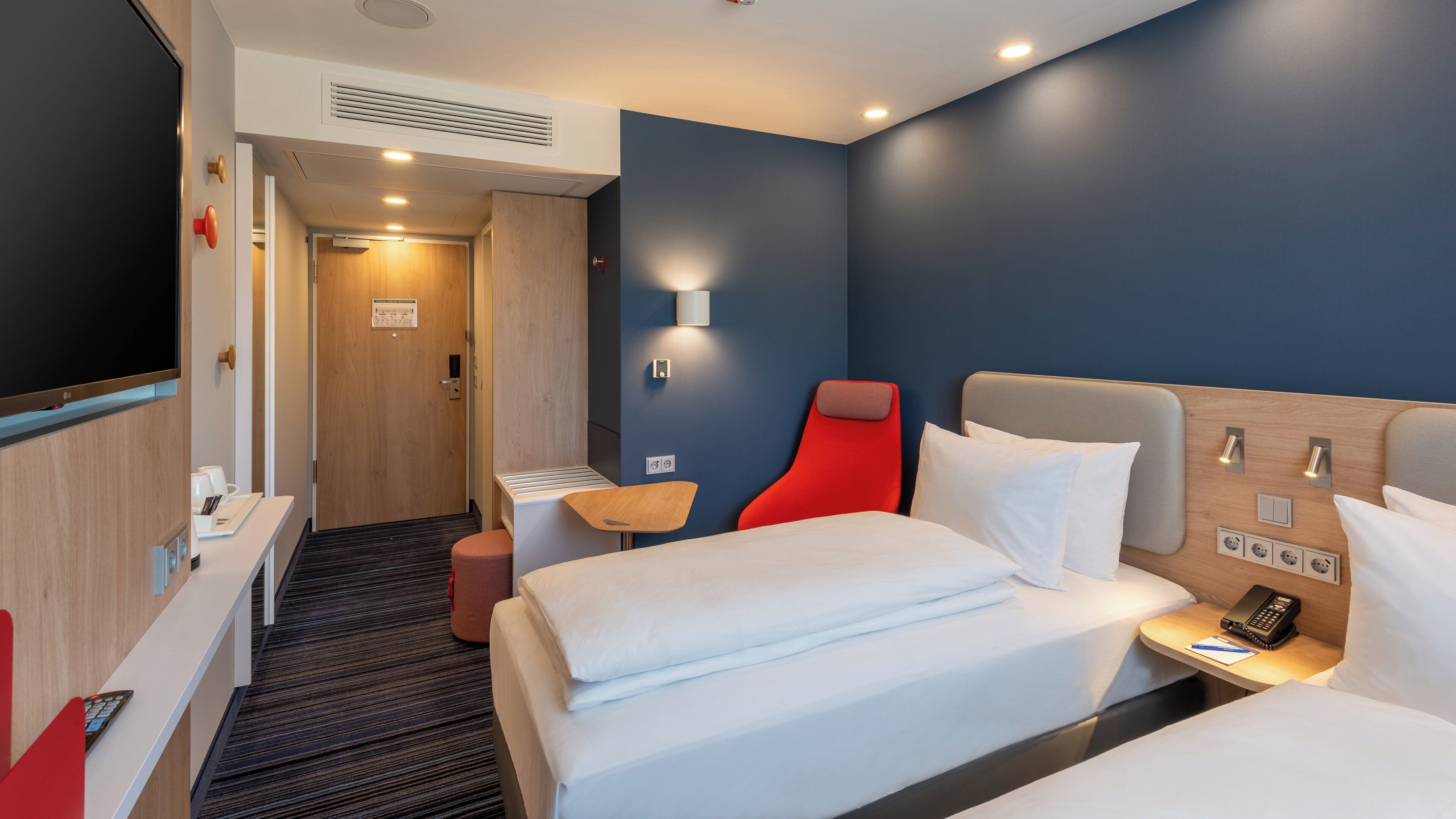 A hotel room with a bed, bedside table, a red chair, a desk with a computer, and a television.