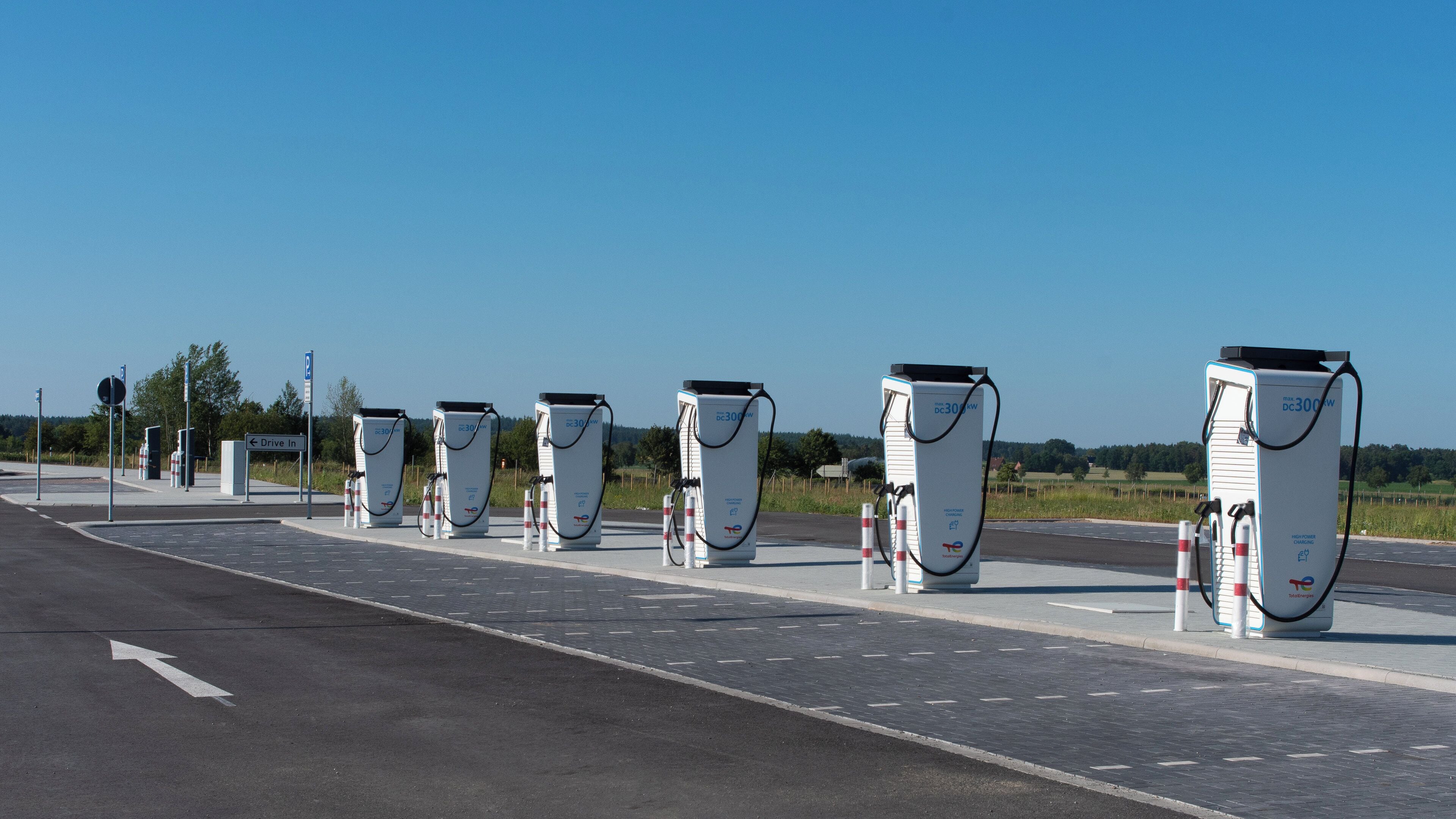 A row of electric vehicle charging stations along a road with directional signs and a clear sky.