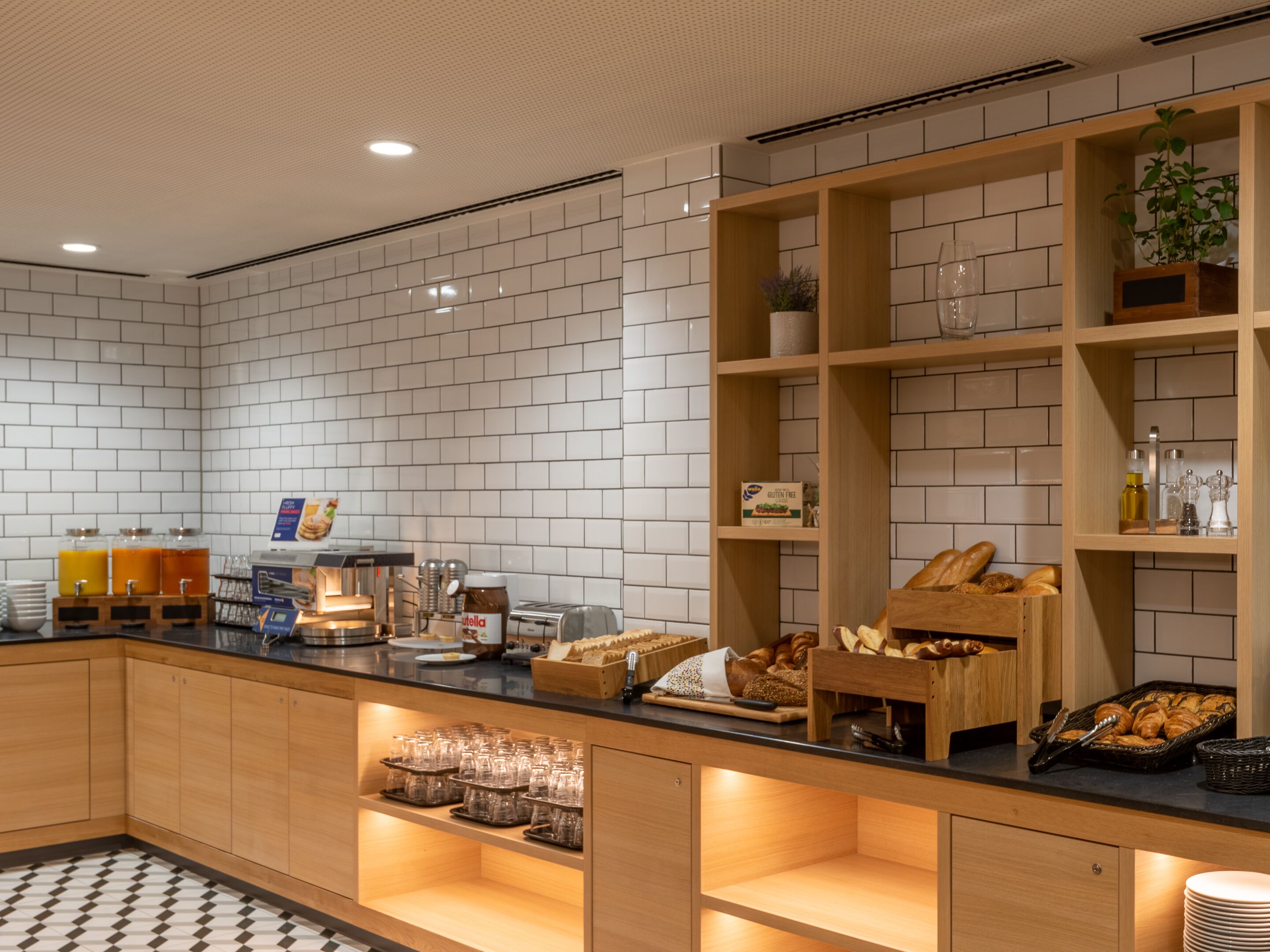 A modern kitchen with white tiled walls, wooden cabinets, and a black countertop. There are glass jars, a coffee machine, and a bread display.
