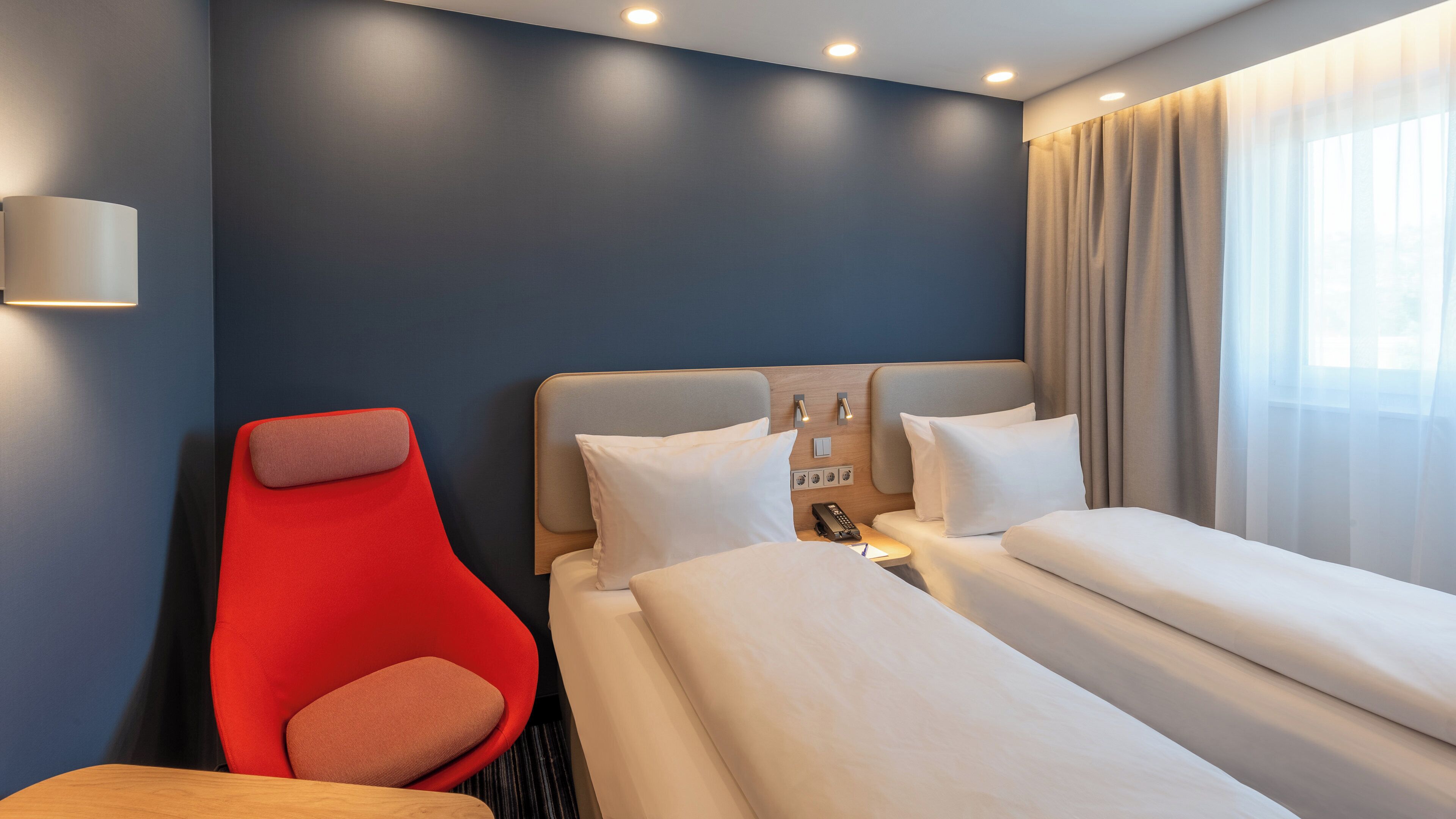 Una habitación de hotel con una cama, una silla roja, una mesita de noche, una lámpara y una ventana con cortinas.