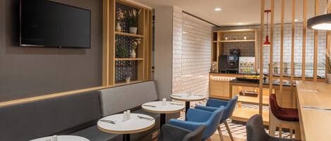 Un restaurante moderno con una televisión en la pared, con disposición de mesas que incluye un banco y mesas redondas, y un área de cocina visible a través de una partición.