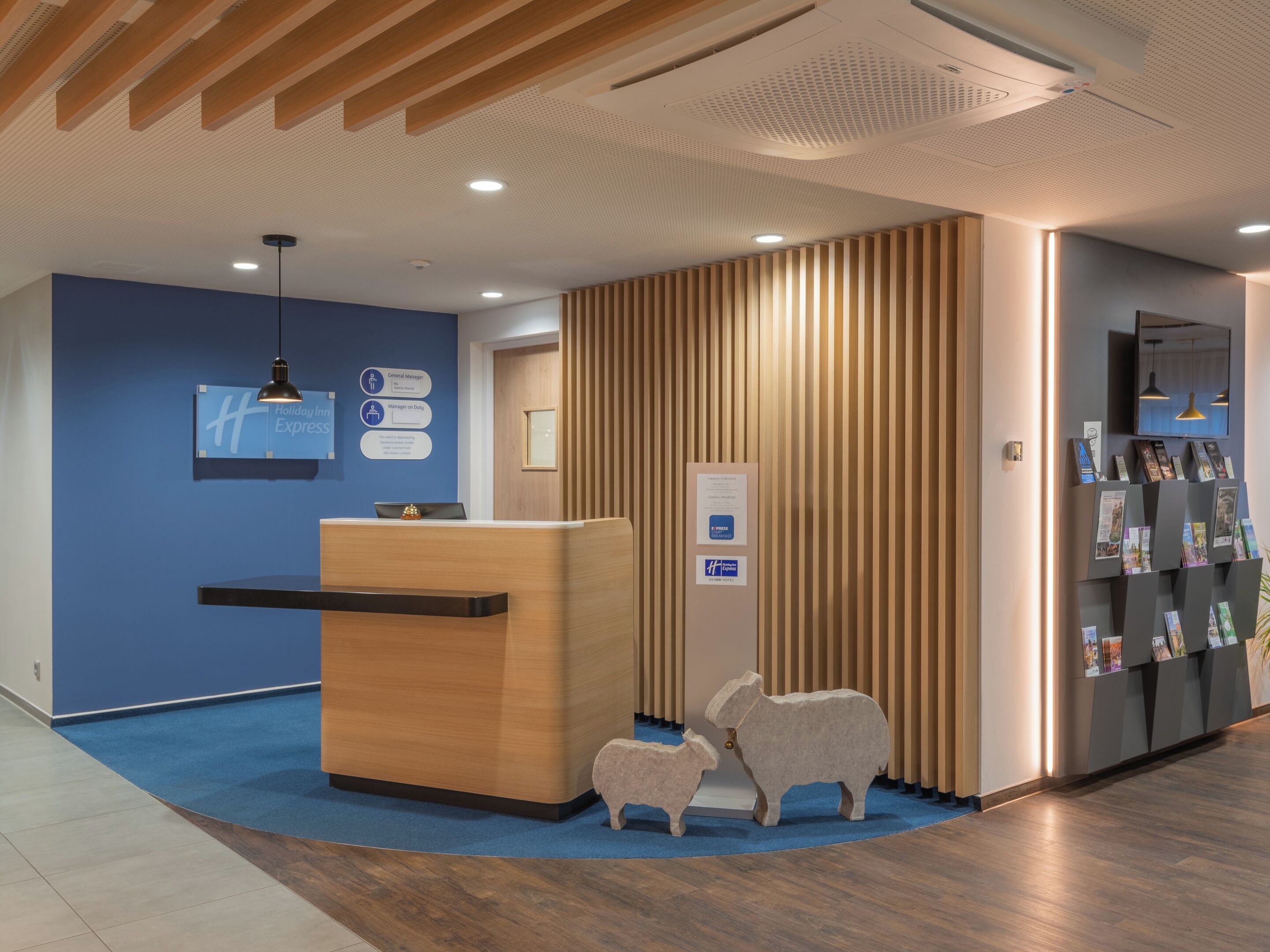 Un área de recepción con un mostrador de madera, alfombra azul y un panel informativo en la pared.