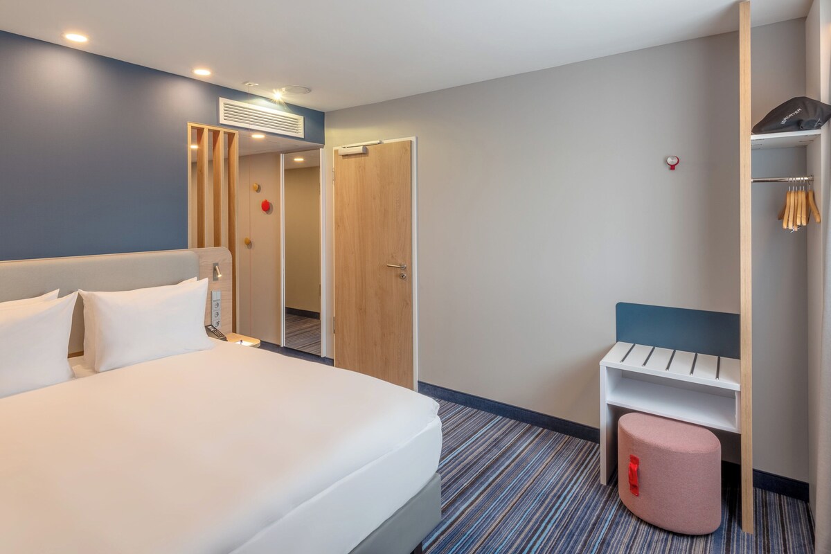 A hotel room with a bed, a bedside table, a wardrobe, and a seating area with a bench.