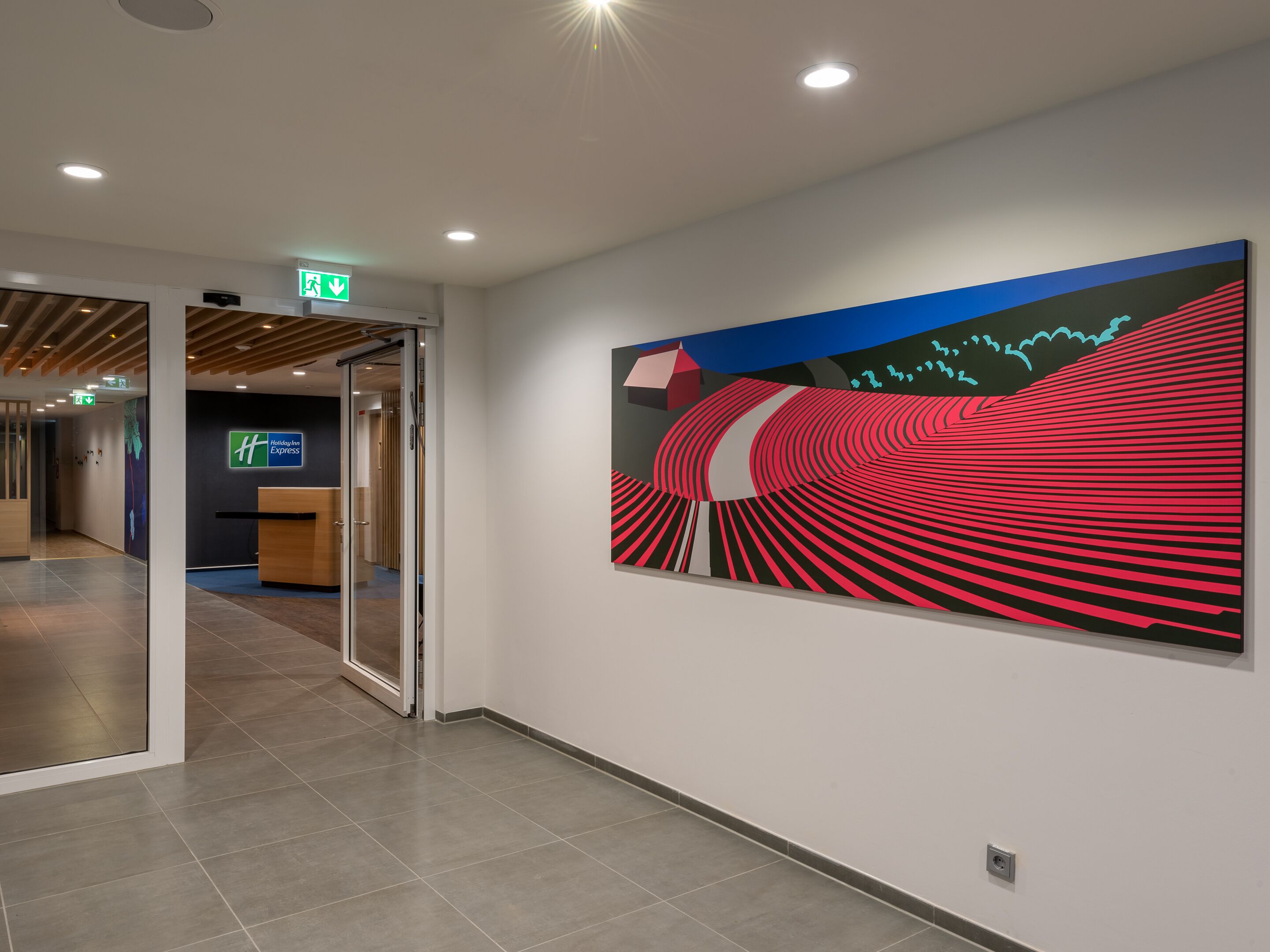 En modern hotellobby med en receptionsdisk, en entré med glasdörr och en stor, abstrakt målning på väggen.