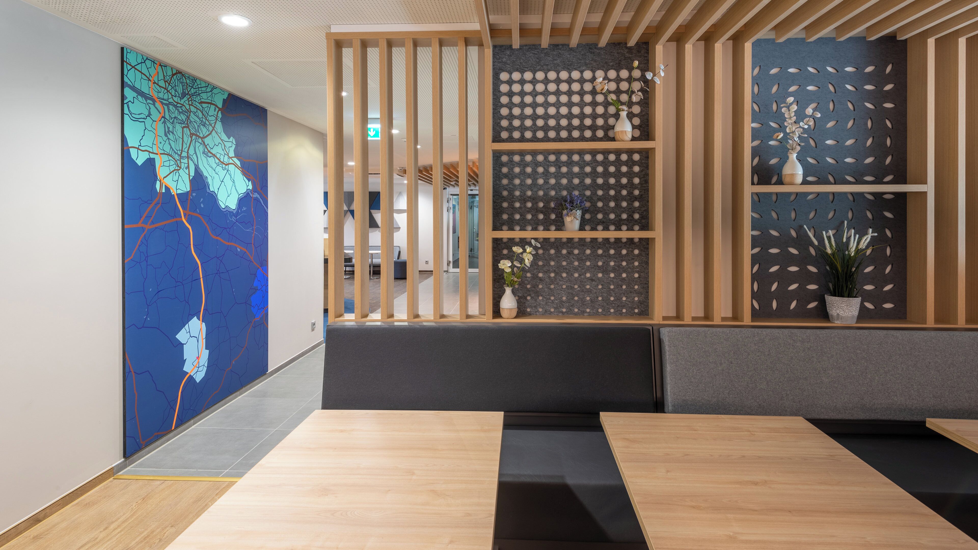 Un interior moderno con una mesa de madera, asientos acolchados grises y una pared decorativa con patrones circulares y plantas.