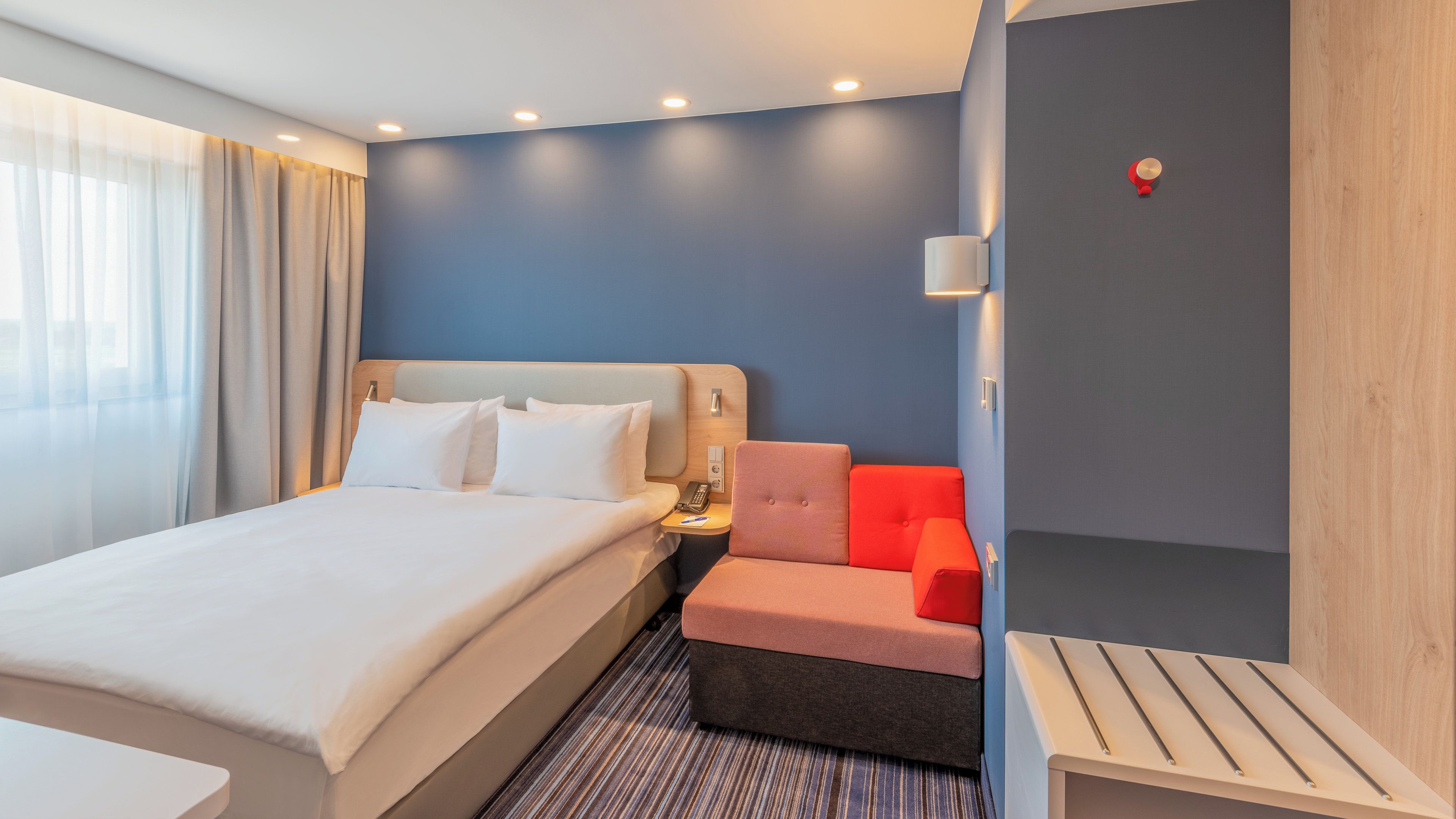 Ein Hotelzimmer mit Bett, Nachttisch, Lampe und einem roten Sessel.