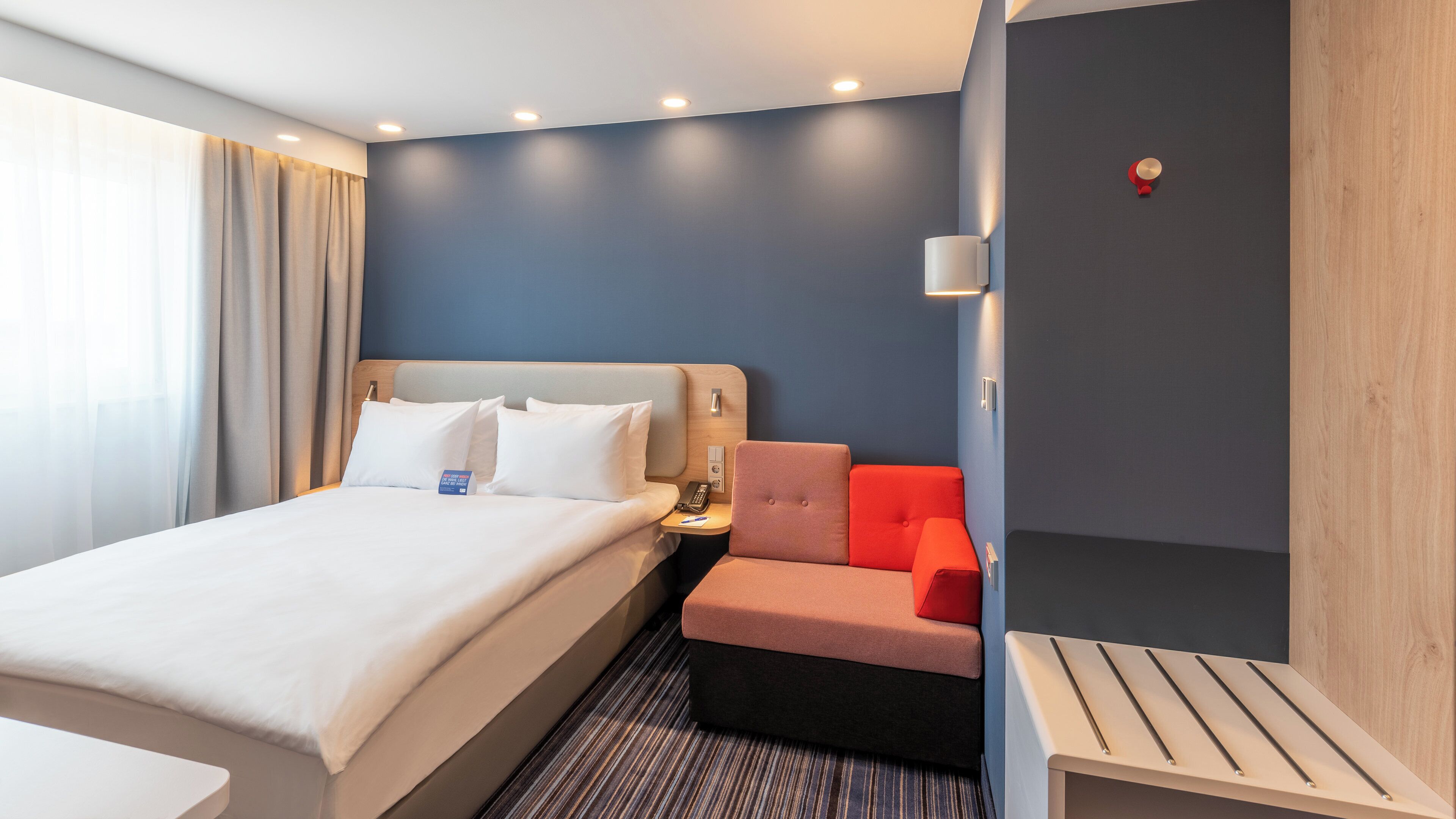 A hotel room with a bed, bedside table, nightstand, and a seating area with a chair and ottoman.