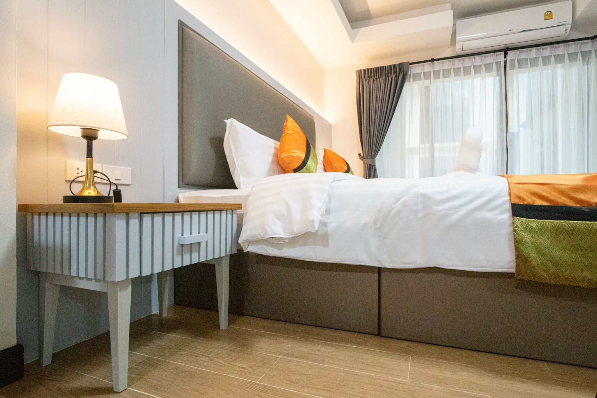 A hotel room with a bed, a bedside table with a lamp, a headboard, and a window with curtains.