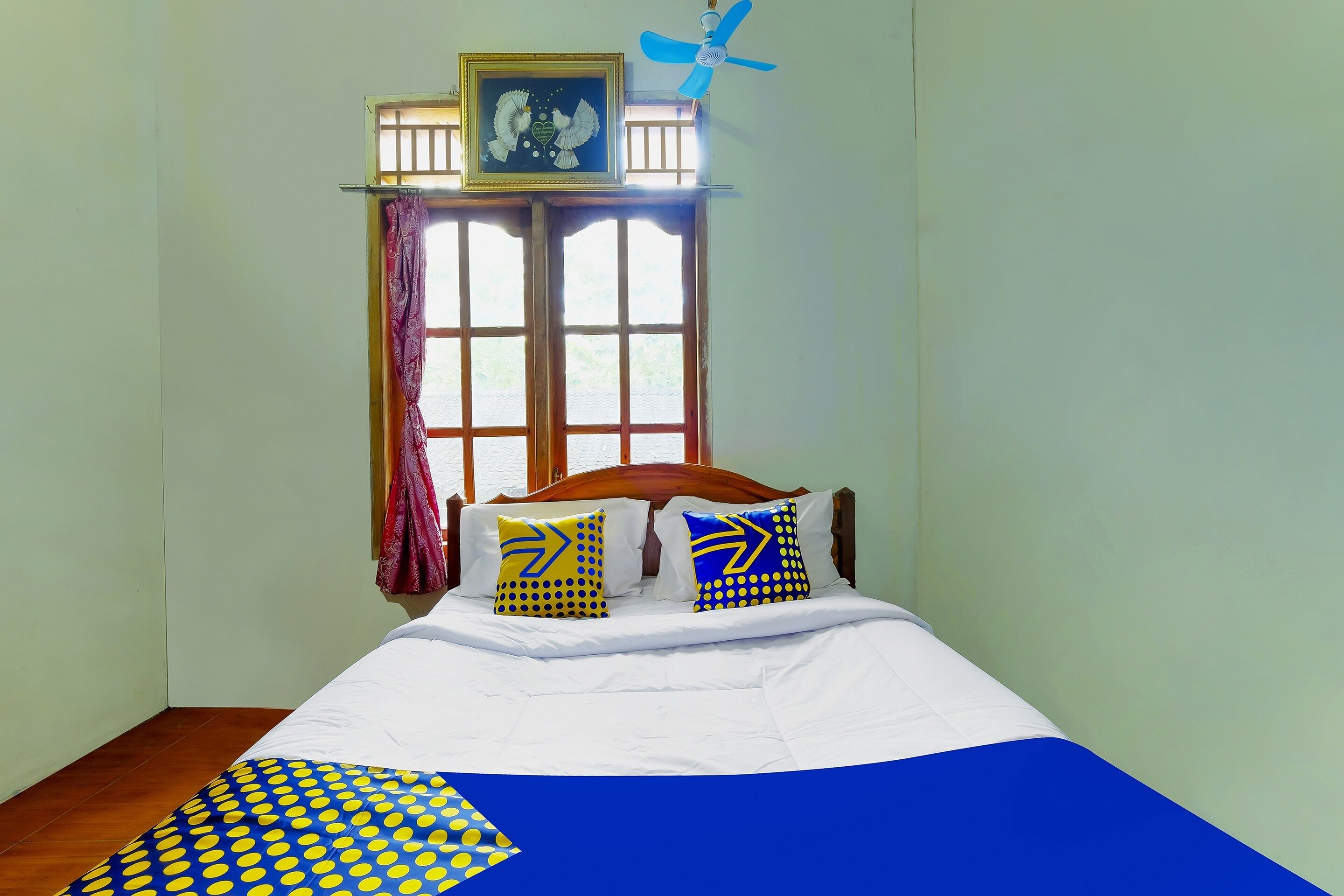 A bed with white bedding and two pillows with blue and yellow patterns, a wooden headboard, a window with curtains, and a ceiling fan.