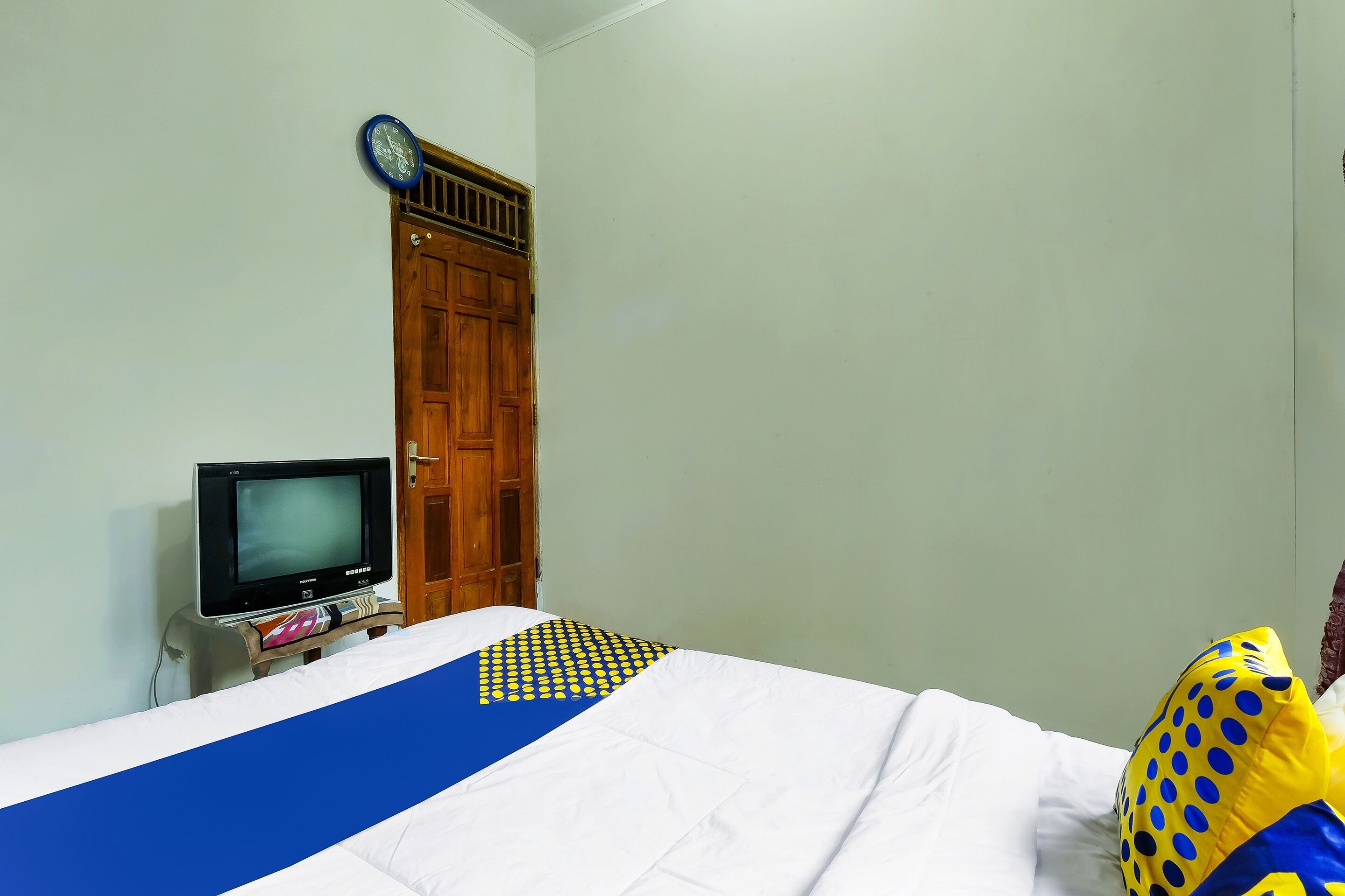 A room with a bed, a television on a stand, a wooden door, and a wall clock.