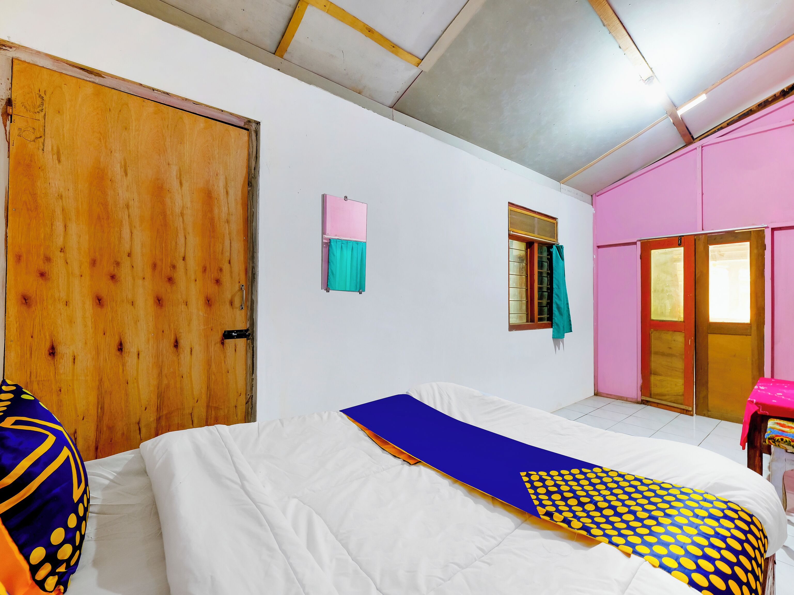 Quarto com porta de madeira, janela com cortina, cama com cobertor azul e amarelo, e parede rosa.