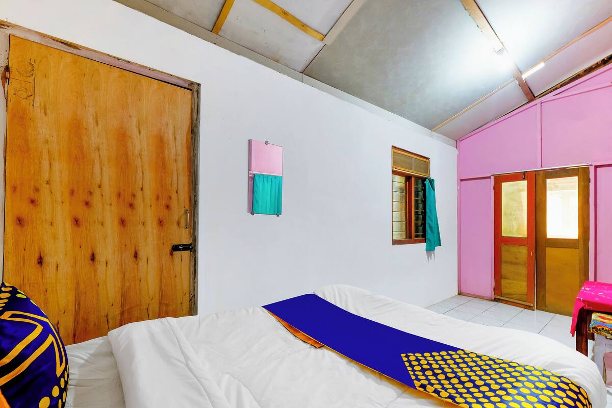 Une chambre avec une porte en bois, une fenêtre avec un rideau, un lit recouvert d’une couverture bleue et jaune, et un mur rose.