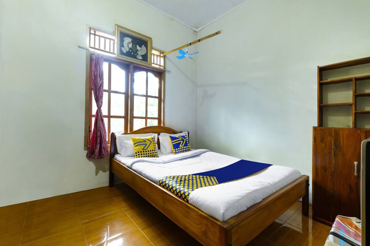 Une chambre à coucher bien rangée, avec un lit en bois, une fenêtre avec des rideaux, un ventilateur de plafond et une armoire en bois.