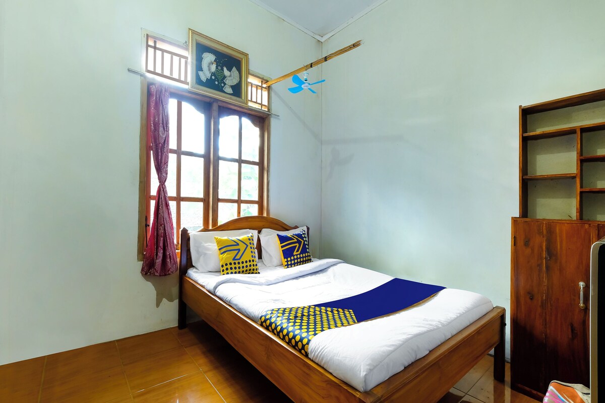 Ein Einzelbett mit Holzrahmen, ein Fenster mit Vorhängen, ein Deckenventilator und ein Holzschrank.