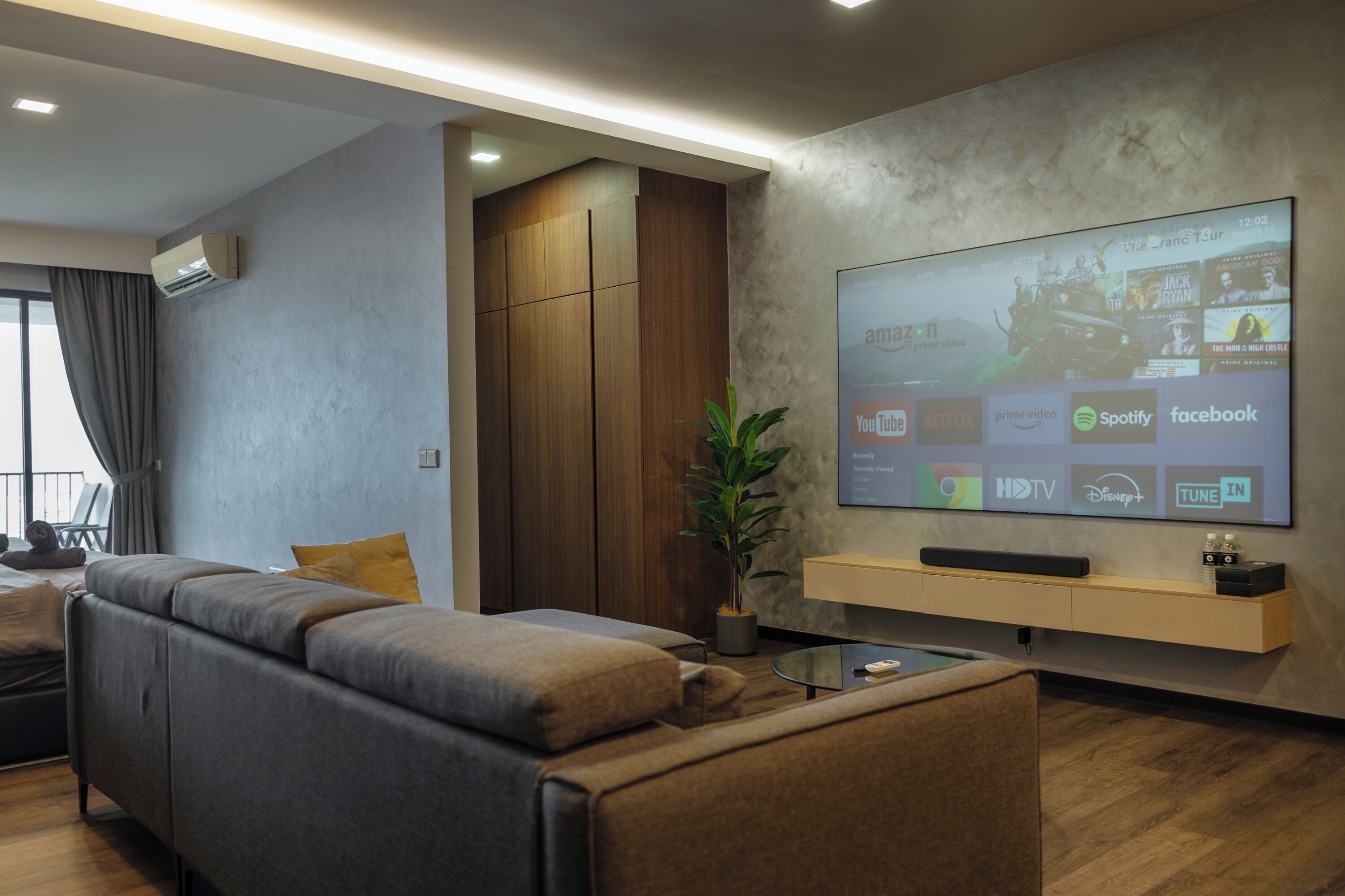A modern living room with a large flat-screen TV mounted on the wall, a comfortable sofa, a coffee table, and a potted plant.