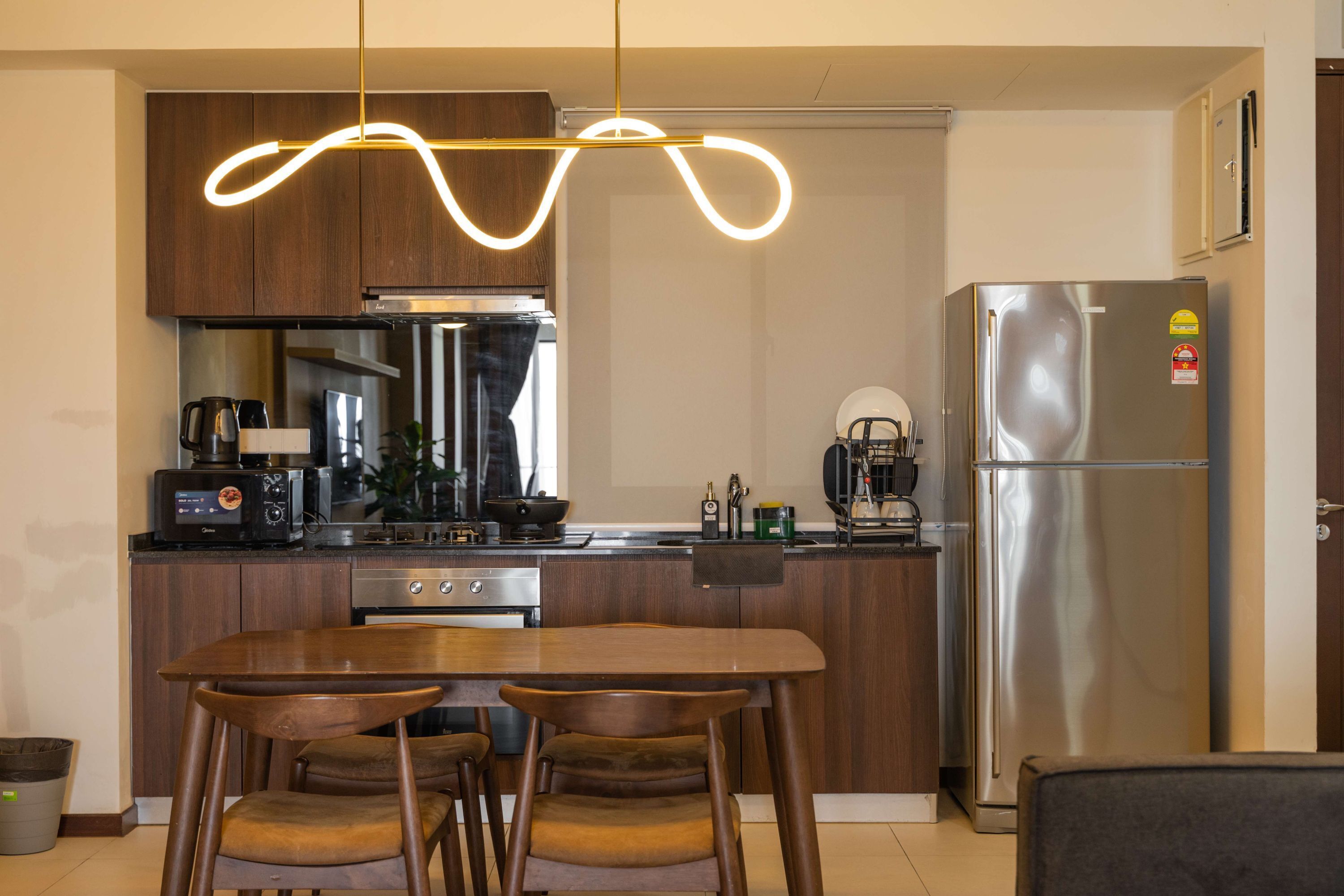 Une cuisine moderne avec des meubles en bois, un réfrigérateur en acier inoxydable, une table à manger avec des chaises et un luminaire suspendu original.
